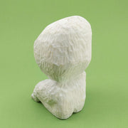Back view. Whittled wooden sculpture of a simplistically shaped white creature, sitting with its hands on its knees. It has a large head to its small body, with large black oval shaped eyes and an open black mouth.