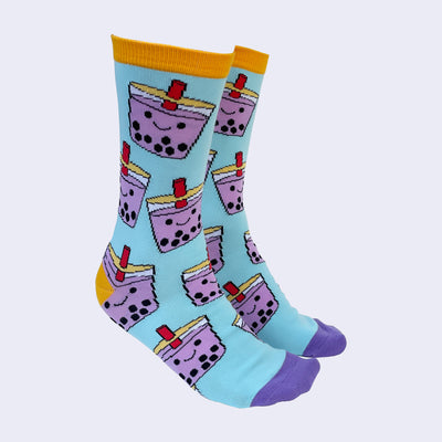 Side view of a model's feet wearing chubby cartoon boba socks. The heel area and trim are a marigold color. The toe area is bright purple.