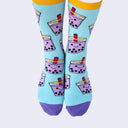 Front view of a model's feet wearing chubby cartoon boba socks. The smiling purple cups are filled with black boba balls. A red straw pokes out.