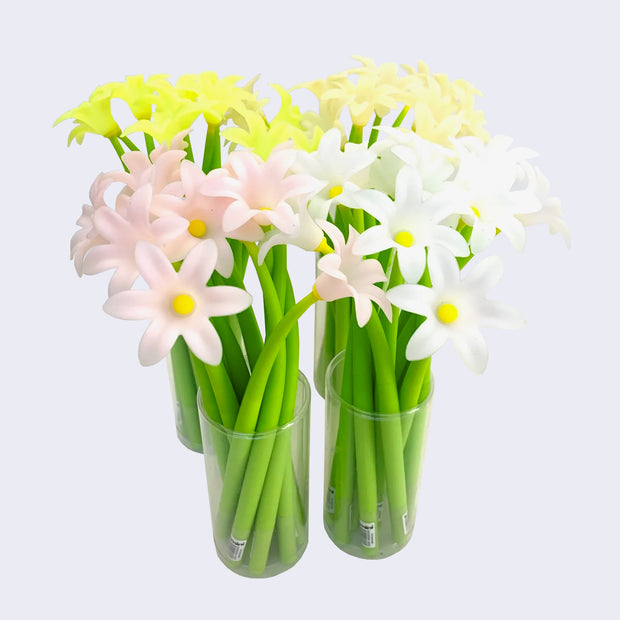 Four clear cups holding green rubber pens with pink, yellow, white and light yellow lily flowers as pen toppers.