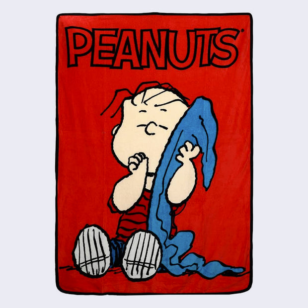 Spread out red plush blanket with "Peanuts" written in logo font at the top. Linus sits, holding his blue blanket and with his thumb in his mouth. Image is large in the center of the blanket.