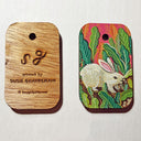Colorful painting on wooden rectangle token with rounded edges and a hole in the upper middle. Can be seen front and back. The back side of the wood has "SG artwork by Susie Ghahremani" burned into it.