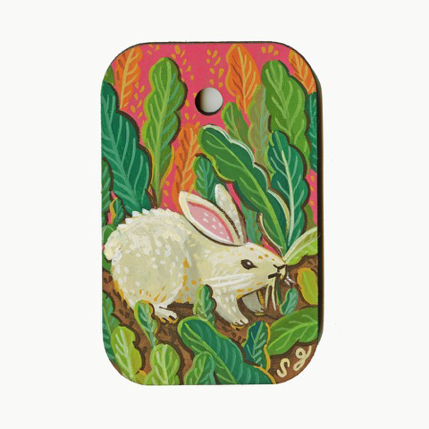 Colorful painting on wooden rectangle token with rounded edges and a hole in the upper middle. A white bunny eats on tall green leaves in dirt, presumably a garden. Background is pink with yellow accents.