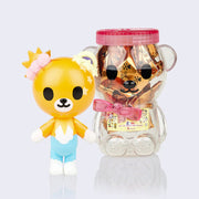 Vinyl cartoon bear figure, standing in front of clear honey bear inspired container, with a pink screw on lid and bow around its neck. Bear is orange with blue pants and white paws, its left ear is a sun and its left ear is a moon.