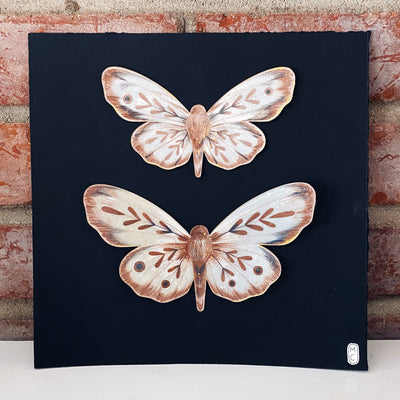 Two brown moths made out of paper, splayed out flat with the smaller of the two on top. They are on black paper.