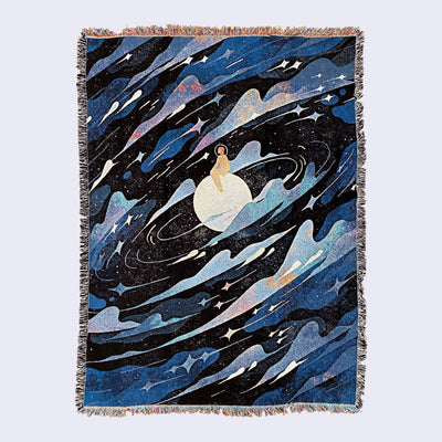 Large tapestry blanket with intentional frayed edges, rainbow on top and bottom and black and white on the sides. Tapestry design is of a figure in an orange space suit and helmet, sitting on a moon. All around them are blue and white galactic swirls, lots of clouds and stars. 