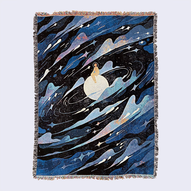 Large tapestry blanket with intentional frayed edges, rainbow on top and bottom and black and white on the sides. Tapestry design is of a figure in an orange space suit and helmet, sitting on a moon. All around them are blue and white galactic swirls, lots of clouds and stars. 