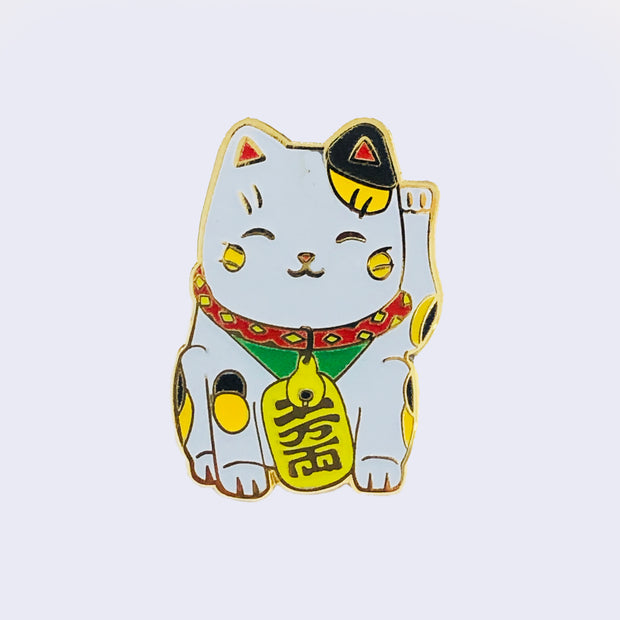 Enamel pin of a happy maneki cat, with yellow and black spots. It has a red, yellow, and green collar and tag with Japanese script hanging from its neck. One paw is up.