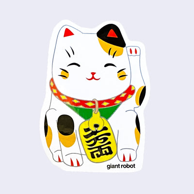 White cut out sticker of white Japanese Lucky Cat with brown and black spots, with a happy expression and one arm raised. A large gold tag is around its neck and "Giant Robot" is written in small black font near its right foot.