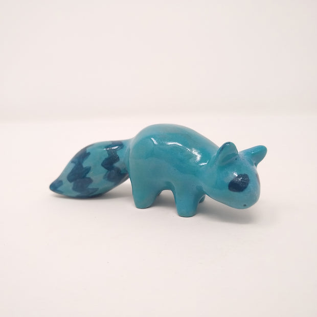 Small blue ceramic raccoon standing on all fours, with a fluffy zig zag striped tail.