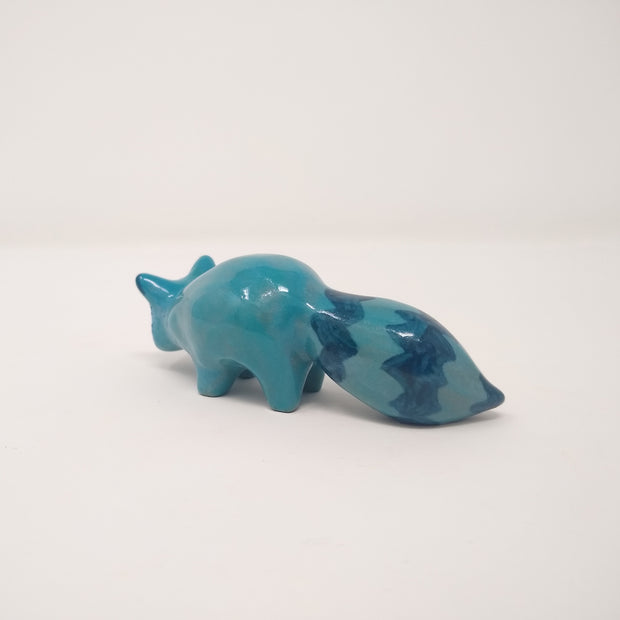 Small blue ceramic raccoon standing on all fours, with a fluffy zig zag striped tail.