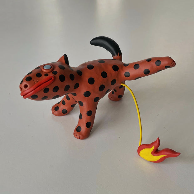 Clay sculpture of a deep orange dog with black polka dots. Its face is simplified in an abstract way, with blue eyes and red mouth. It lifts one of its legs and a stream of yellow comes out of it, leading to a small orange and yellow flame.