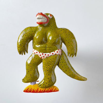 A semi-flat wall hanging sculpture of a smooth, buff Godzilla like creature with large red lips and many teeth. Its skin is olive green with yellow and black polka dots, wearing a white with red polka dot bikini bottom. It stands with one foot atop a tiny tank, as if mid smash.