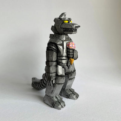 A carved wooden sculpture of a mechanized Godzilla, painted silver and black with bright yellow eyes, holding a strawberry ice cream cone with sprinkles.