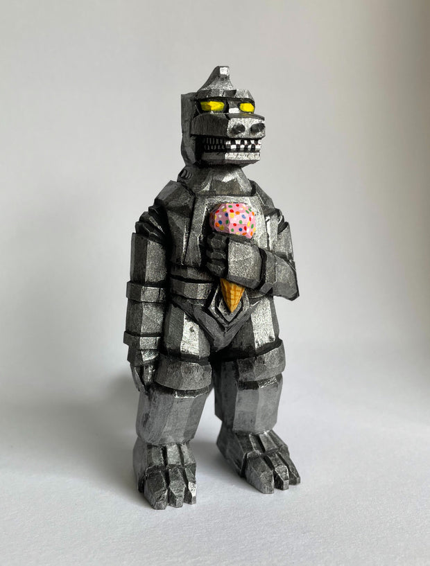 Front view of a carved wooden sculpture of a mechanized Godzilla, painted silver and black with bright yellow eyes, holding a strawberry ice cream cone with sprinkles.
