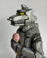 Close up view of a carved wooden sculpture of a mechanized Godzilla, painted silver and black with bright yellow eyes, holding a strawberry ice cream cone with sprinkles.