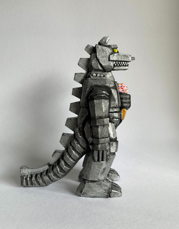 Side view of a carved wooden sculpture of a mechanized Godzilla, painted silver and black with bright yellow eyes, holding a strawberry ice cream cone with sprinkles.