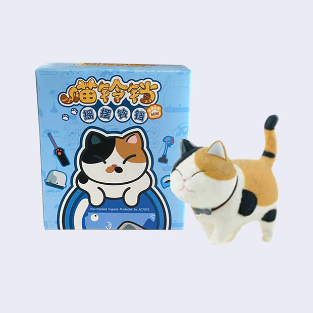 Vinyl figure of a chubby calico cat, with a closed eye expression of contentment and a black collar with silver accent. It stands in front of a display box, blue with Japanese script and illustration of a smiling cat hanging over a tin can.