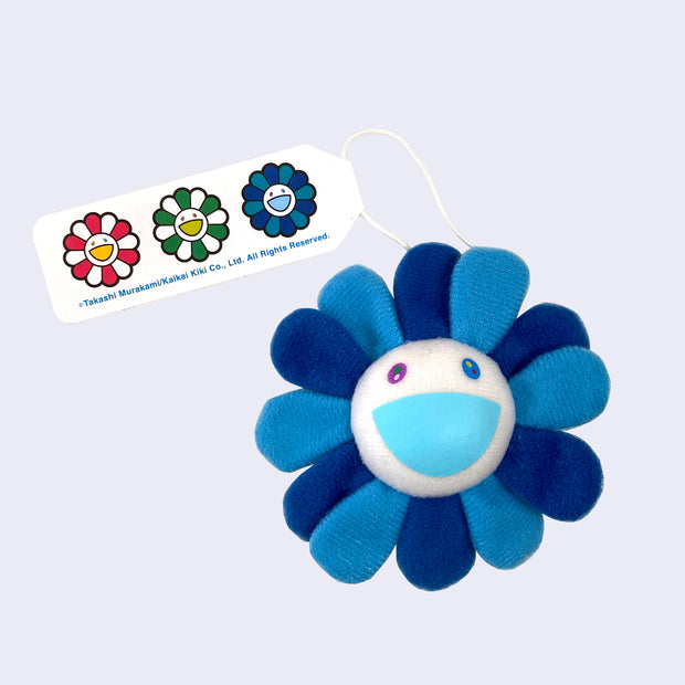 A Murakami flower with a cheerful expression, white face with a light blue smile and alternating dark and light blue petals, with a hanging tag attached.
