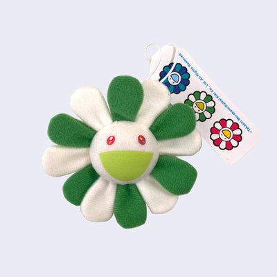 A Murakami flower with a cheerful expression, white face with a light green smile and alternating dark and light green petals, with a hanging tag attached.
