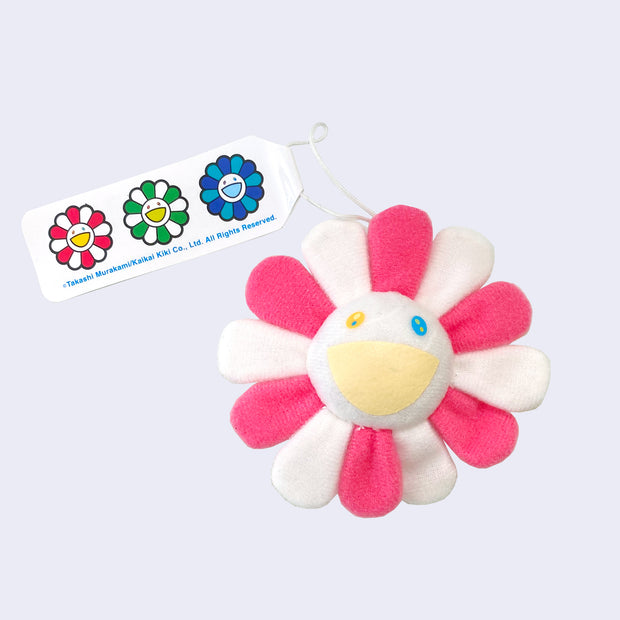 A Murakami flower with a cheerful expression, white face with a light yellow smile and alternating pink and white petals, with a hanging tag attached.