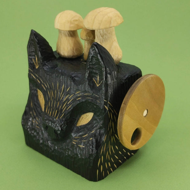 Side angle of a wooden, block shaped black wolf head with small details carved exposing the natural wood, there are 3 wooden mushrooms atop its head and a turnable disc handle on the side.