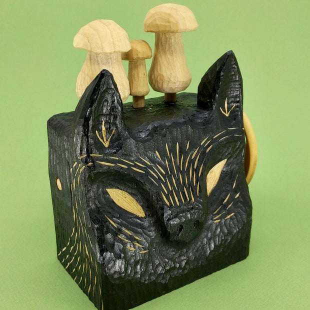  A wooden, block shaped black wolf head with small details carved exposing the natural wood, there are 3 wooden mushrooms atop its head and a turnable disc handle on the side.
