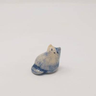 Small ceramic sculpture of a little blue and cream colored cat, looking up but curled into itself. It has a simplistic painted on face with whiskers.