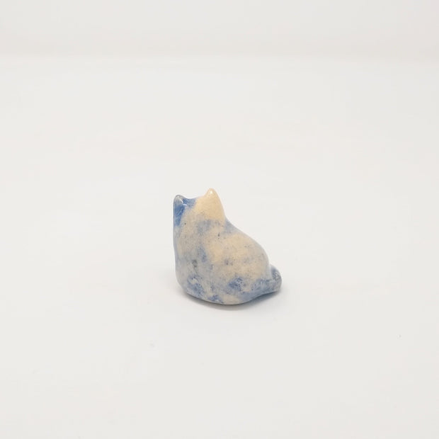 Small ceramic sculpture of a little blue and cream colored cat, looking up but curled into itself. 