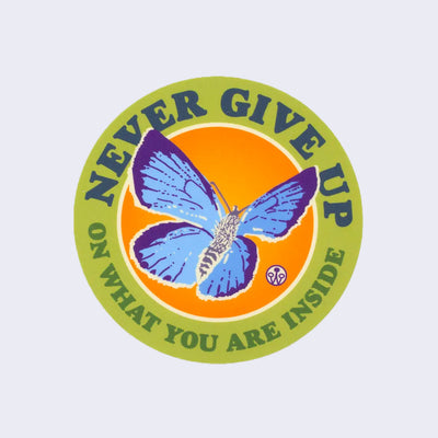 Circular sticker with lime green border and orange background. An illustrated blue and purple butterfly flies with "Never give up on what you are inside" written in bold font around the borders.