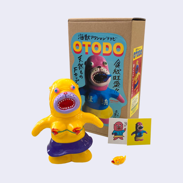 Bright yellow painted seal-like soft vinyl figure, standing with colorful eyes, an open mouth and wearing a shrimp shaped bikini and blue skirt. A small plastic yellow with orange polka dot fish lays at its feet.