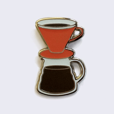 Gif of enamel pin of a pour over coffee set, with an orange dripper and full pot of black coffee. One image is of pin in light, the other is of pin glowing in the dark.