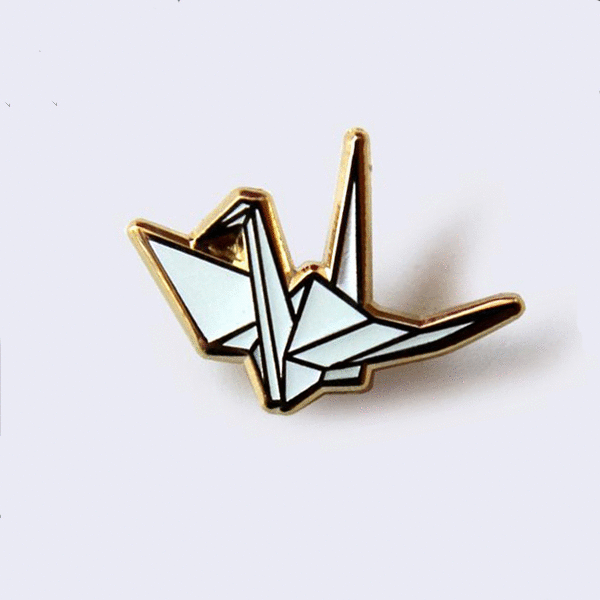Gif of an enamel origami crane pin. One image is of the pin in light and the other is of the pin glowing in the dark.