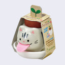 Small round ceramic gray cat planter with a large pink fabric tongue. It sits in a small round dish and is encased in cardboard packaging that reads "Peropon"