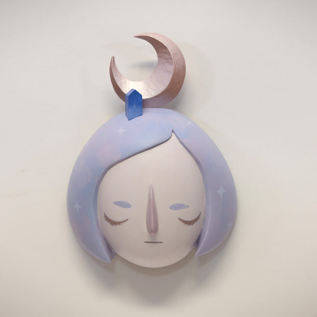 Painted wooden sculpture of a stylized face with closed eyes and a purple bob haircut with a side part. Atop her head is a small blue building and a metallic crescent moon. 