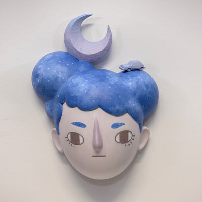 Painted wooden sculpture of a stylized face, with lumpy blue galaxy patterned hair. Atop their head is a purple crescent moon and a small purple car.