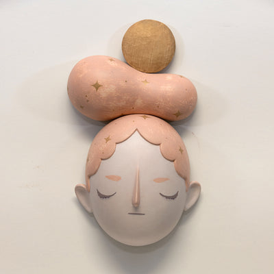 Painted wooden sculpture of a stylized closed eye face with light pink hair with gold sparkles. Atop the head is a pink gourd shape with gold sparks with a natural wooden sphere above. 