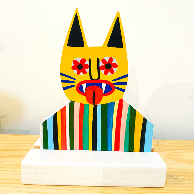 Die cut painted wooden sculpture of a yellow cat with pointy ears, tongue out and flowers for pupils. It wears a striped shirt and is only visible from the chest up.
