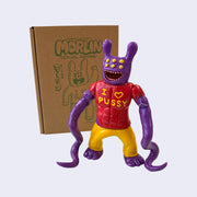Purple painted soft vinyl figure, with 6 yellow eyes a wide open mouth smile and two bunny like ears. The figure is muscular and wears a red shirt that says "I heart pussy" with two long tentacles as arms.
