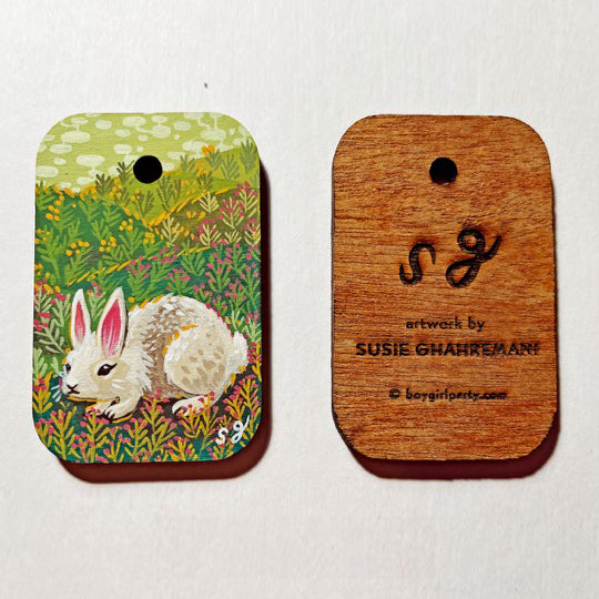 Colorful painting on wooden rectangle token with rounded edges and a hole in the upper middle. Can be seen front and back. The back side of the wood has "SG artwork by Susie Ghahremani" burned into it.