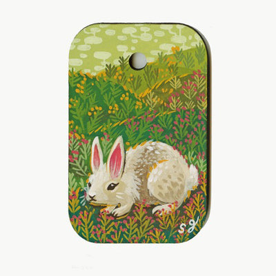 Colorful painting on wooden rectangle token with rounded edges and a hole in the upper middle. A white fluffy rabbit with pink ears sits in a setting of rolling hills, with many brush flowers blossoming atop the hills. Background is light green sky with white bubble pattern clouds.