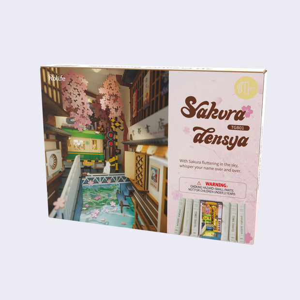 Rectangle box packaging that reads "Sakura Densya" and includes a close up photo of the assembled model kit, a Japanese alley way with a body of water, a tram going through and cherry blossom trees.