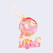 Semi transparent sculpted figure of a small person sitting with their hands at their side, legs out in front of them and wearing a scuba diving helmet with a feather coming out of the top. Figure is primarily pink with orange ombre elements.