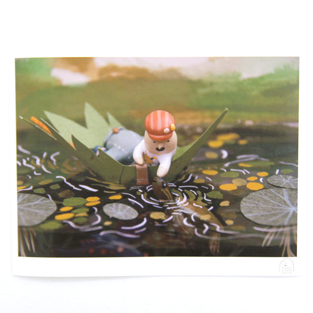 Photograph of clay and paper diorama. A person floats on a body of water in a leaf made into a boat. They reach out from their boat to pet a turtle that appears out of the water.
