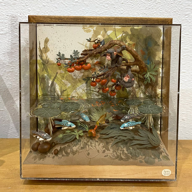 Diorama like sculptural piece encased in a glass box with a wooden mount framing. Elaborate scene of two monkeys hanging onto the branch of a persimmon tree, looking at the water below them, filled with water plants and various colored fish.