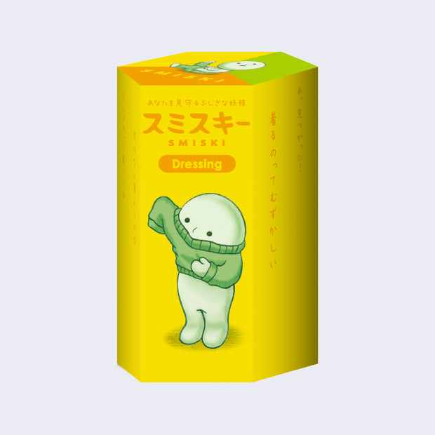 Yellow cylindric blind box packaging, with Japanese script and text that reads "Smiski Dressing" with an illustration of a simple, light green character pulling on a green sweater.