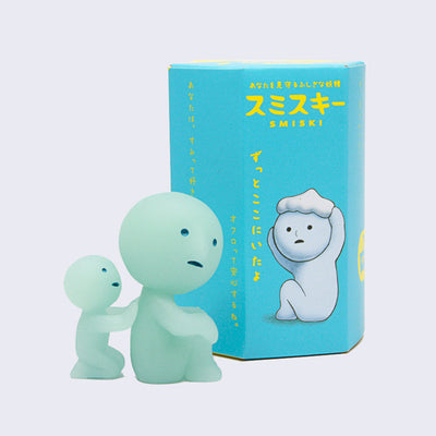 Simplified blue character with a solemn expression, hugging its knees with a smaller character touching its back behind. Behind is a blue display box, with Japanese script and "Smiski" written in yellow.