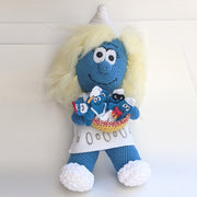 A crocheted plush sculpture of a smiling Smurfette, holding a bowl filled with various sewn Smurfs that appear flat like gingerbread cookies.