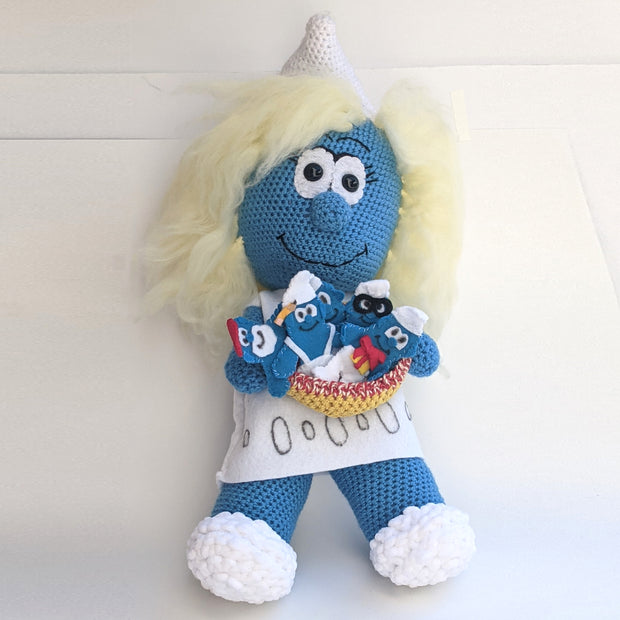 A crocheted plush sculpture of a smiling Smurfette, holding a bowl filled with various sewn Smurfs that appear flat like gingerbread cookies.