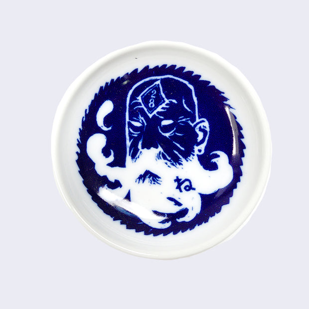Small white ceramic saucer plate featuring a dark blue illustration of an old man with a long whispy beard. On his forehead is a diamond with 28 written on it.